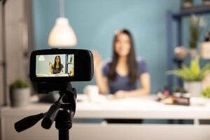 Selective focus on a device screen showing a smiling woman seated at desk, doing a hello hand gesture, vlogging in a studio. Digital camera on tripod records a beauty influencer during a livestream. photo
