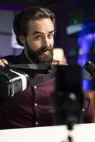Tech professional reviewing VR glasses in neon lit studio, presenting specifications. Man filming virtual reality headset review for technology fans using cellphone on tripod stabilizer photo