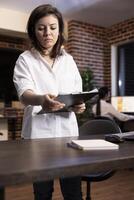 Caucasian female employee stands at her workstation, analyzing documents on a clipboard. She flips through pages, reviewing important information during a late night work session. photo