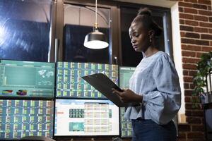 Businesswoman standing with a clipboard, reviewing a financial report and preparing an investment strategy in modern office setting. Black female entrepreneur writing notes based on trading progress. photo