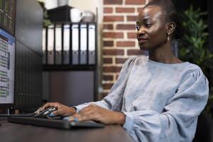 African American trader reviews financial statistics on multiple monitors, studying live stock market data in focused work setting. Black woman at desk, using computer mouse and keyboard in office. photo