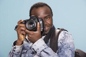 African American male photographer demonstrating the usage of a DSLR. Smiling black man, carrying a backpack, holds a professional camera close to his face as he focuses on his next shot. photo
