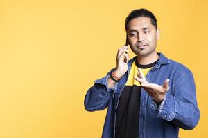 Young adult with man bun discussing with best friend over telephone call, enjoying leisure time in studio. Upbeat guy in casual clothing talking with mate on phone call, studio backdrop. photo
