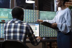 Two traders working late at night in a modern office, collaborating on multiple monitors displaying forex market charts and stock data. African american business partners reviewing stock trading data. photo