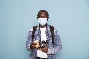 Professional photographer wearing a face mask, holding a DSLR camera and a coffee cup as he stands in blue background studio. Portrait of black man in casual outfit, covering his mouth for protection. photo