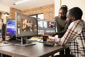 Skilled creative team using multi monitor setup to improve film frame quality in brick wall workspace. Professional male editor working at desk as female teammate assists in enhancing footage visuals. photo