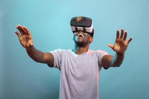 Energetic black adult enjoying VR experience, fully engaged in a futuristic simulation with gaming glasses. Male tech enthusiast immersed in a 3d digital world, curiously reaching out with his hands. photo
