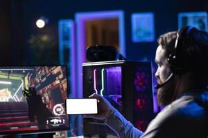 Man watching tutorial on isolated screen mobile hone while playing first person shooter videogame with guns. Gamer learning to play game by looking at online guide on mockup cellphone photo