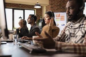 Multiethnic call center agents work at their stations, engaging with remote customers and processing service requests. Computers, coffee cups, and headsets set the scene for a busy office. photo