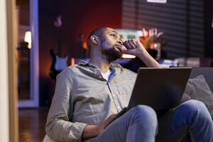 Focused african american guy multitasking on laptop in stylish living space, connected to the internet while managing his freelance work and remote responsibilities. Browsing web laying on sofa. photo