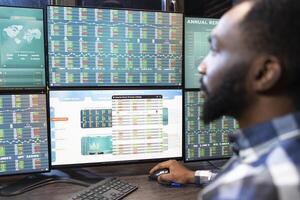 Male financial analyst studying company hedge funds on multiple monitors in brick wall office. Closeup of african american employee focused on stock trading and investment data at his tech savvy desk. photo