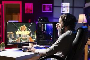 Gamer in neon lit apartment excited after winning level in first person shooter, killing all enemies. Happy man celebrating, manifesting himself after being victorious in videogame photo