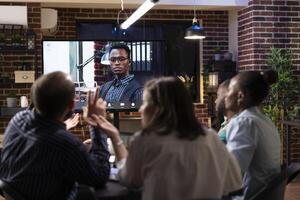 Multiethnic entrepreneurs working late in brick wall office, engaging in online business discussion with coworker on call. Virtual meeting with remote businessman is projected on digital monitor photo