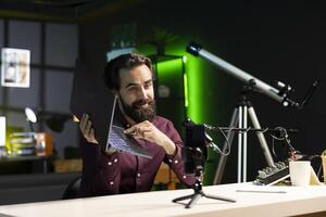 Media star in studio endorsing laptop from brand sponsoring , filming with smartphone on tripod. Tech expert does influencer marketing by promoting notebook, recording with phone photo
