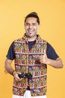 Portrait of overjoyed indian gamer showing thumbs up sign, holding controller, playing videogames. Ecstatic person with gamepad delighted to do gaming, doing approving hand gesture, studio backdrop photo