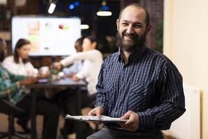 Portrait of caucasian male executive with clipboard, sitting in office, overseeing startup team. The evening setting, brick wall and digital devices emphasize a productive, modern workspace. photo