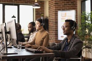 African American man in wheelchair seated at his workstation, wearing headset and using desktop PC. Modern office desk with computer screen, highlighting a sleek and organized business environment. photo