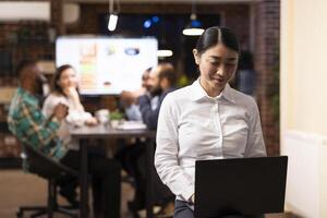 Focused female financial advisor checks reports on her laptop while diverse startup team collaborates in modern office. The workspace reflects teamwork, consulting and business strategy. photo
