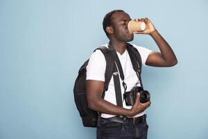 Black photographer in casual clothing carrying backpack and enjoying a hot beverage. African american man with a camera around his neck takes a sip of coffee while standing against isolated background. photo