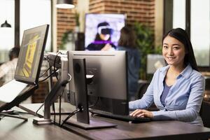 Businesswoman seated in a brick wall workspace, using desktop pc, replying to emails. Asian female creative working at her office desk, typing on a computer keyboard and smiling at the camera. photo