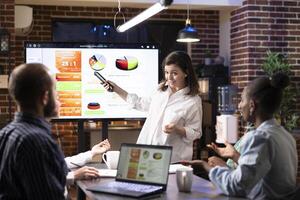 Professional woman explaining business strategies to coworkers using digital monitor. Diverse team attentively reviewing financial charts, collaborating on marketing trends in brick wall workspace. photo