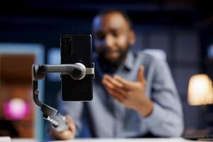 Focus on mobile phone attached to selfie stick used by content creator to do livestream. Enthusiastic online streamer using cellphone to do live broadcast, blurry background photo