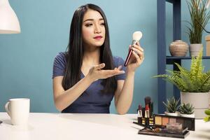 Beauty vlogger explaining makeup tips while holding a brush. Asian woman seated at her desk, gesturing towards the cosmetic tool, making a tutorial and connecting with her online followers. photo