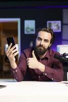 Tech guru reviewing latest phone, giving it thumbs up gesturing while filming internet show in rgb lights studio. Influencer endorsing best smartphone, happy with performance photo
