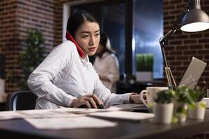 Focused businesswoman multitasking in modern office environment. Dedicated female executive talking on her smartphone, managing documents, and working on her laptop, balancing work tasks effortlessly. photo