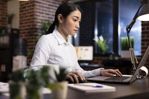 Young businesswoman in professional setting, seated at her desk, using a laptop to prepare a project presentation. The modern brick wall design adds a stylish and productive atmosphere to the office. photo