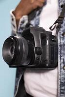 Selective focus on camera worn by black individual in casual outfit, emphasizing design of device. Closeup of DSLR hanging over white shirt and denim jacket, focusing on details of photography gear. photo