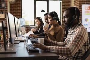 Team of diverse telemarketers sits at their desks, assisting remote clients and managing business inquiries. Headsets, coffee, and computers highlight the dynamic customer support workspace. photo