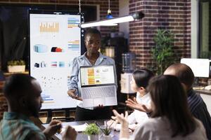 Focused female manager presents business analytics on laptop during night shift meeting. Multiethnic coworkers engage in discussion, analyzing financial charts on digital devices in modern workspace. photo