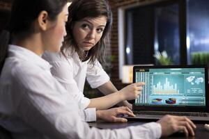 Two female colleagues collaborating on business analytics, reviewing data and discussing strategies on a laptop. They analyze financial information and plan tasks ahead in a professional workspace. photo