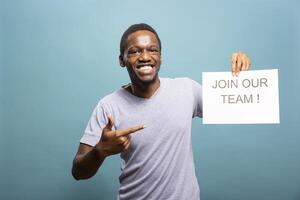 Cheerful male recruiter holding and pointing to a paper in his hand, calling out for people to join his team. African american man gesturing to a hiring sign and smiling at camera. photo