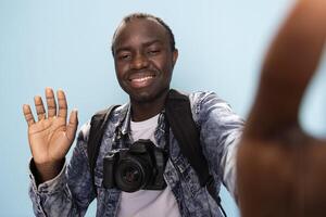 African American man with DSLR around his neck, makes hello gesture, recording a for his vlogging channel. Black male photographer smiles and waves at the camera in studio with blue background. photo
