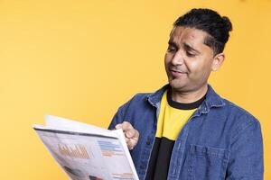 Indian young guy browsing though financial audit report paperwork, feeling overworked and confused of the balance numbers. Accountant person standing against studio background. photo
