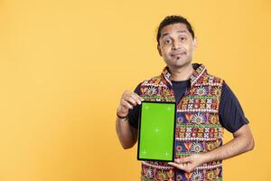Portrait of influencer smiling while presenting green screen tablet, studio background. Upbeat indian content creator holding chroma key device used for advertising brands photo