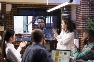 Diverse startup team working late in brick wall office, actively engaging in hybrid meeting with black businessman. Caucasian manager presents while others listen, with call on digital screen. photo