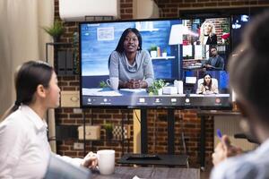 Entrepreneurs in startup office participating in videoconference to discuss project planning. Multiethnic coworkers use digital monitor to collaborate on business goals and marketing tactics remotely. photo