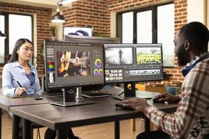 Black media creator operates editing software, merging clips and audio in a diverse production department office. African american man seated at shared creative workstation, working on projects. photo