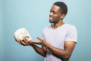 African American man curiously admiring a human skull, holding it gently against an isolated background. Black male model standing and gesturing to a skeletal head in a simple studio setting. photo