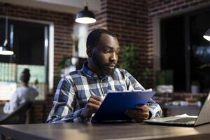 African american male manager sits at his desk, jotting down progress updates on a clipboard for company hedge fund. Financial advisor taking notes as he analyzes stock trading risks in the office. photo