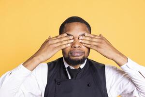 Restaurant employee doing the three wise monkeys symbol on camera, covering his eyes to see no evil as a proverbial metaphor. Wise butler in apron shows symbolic concept in studio. photo