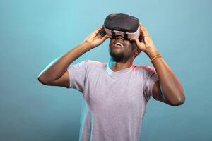 Modern adult playing an interactive 3D game against a blue background, fully immersed in virtual entertainment. Black male model touching his vr headset, looking up, exploring a futuristic simulation. photo