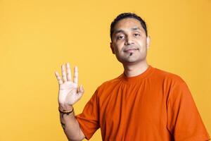 Portrait of lively overjoyed guy waving at a person against orange background, greeting people and showing his smile on camera. Upbeat friendly indian man being overjoyed in studio. photo