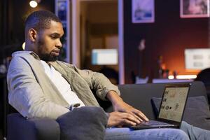 African american specialist working from home using financial graphs analysis, reviewing statistical data to compare supply and demand numbers. Examining business charts on his laptop. photo