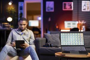 Black man day trading from home on laptop following stock market charts and financial graphs. Remote work setup shows an investor tracking prices, trends and data for online investment decisions. photo