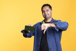 Confident guy doing the thumbs down sign next to mockup on mobile phone, creating an advertisement for a promotion against studio background. Indian man does hand gesture. photo