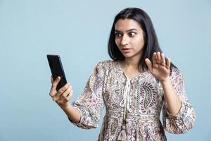 Relaxed woman excited to chat with friend over online call on smartphone, doing friendly gestures and waving. Joyous indian girl talking in teleconference using mobile phone, studio background. photo